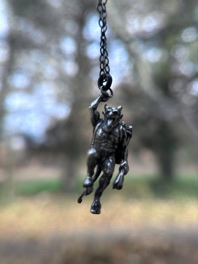 May include: A silver-toned pendant necklace featuring a detailed depiction of a horned, humanoid figure with a tail. The pendant is suspended from a delicate chain, showcasing the intricate design of the mythological creature.