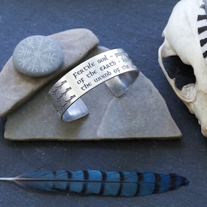 May include: Silver bracelet with an engraved quote: "Fertile soil - pulse of the earth - heart of the womb of the world". The bracelet is on a gray stone with a small stone with a symbol engraved on it. A blue bird feather is on a gray surface.