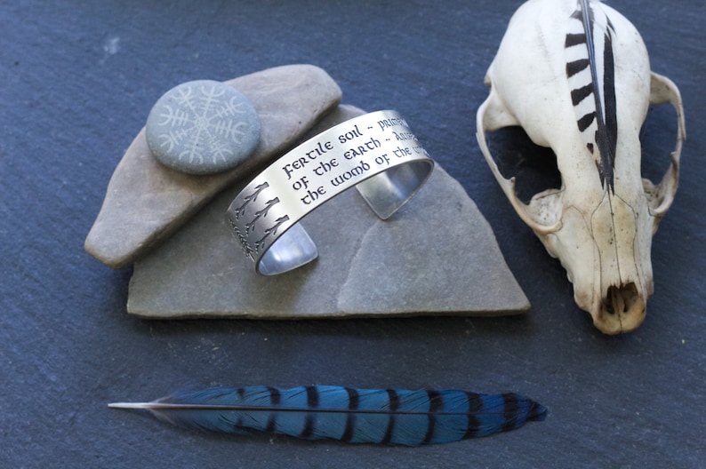 May include: Silver bracelet with an engraved quote: "Fertile soil - pulse of the earth - heart of the womb of the world". The bracelet is on a gray stone with a small stone with a symbol engraved on it. A blue bird feather is on a gray surface.