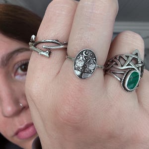 May include: A hand displays four silver rings. One ring is a twig design, another features two figures under a crescent moon, and a third has a green stone. The fourth ring is a Celtic knot design.