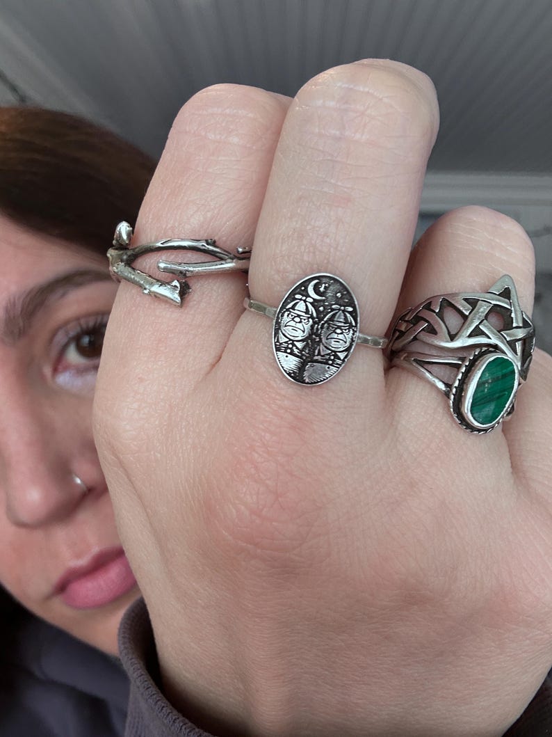 May include: A hand displays four silver rings. One ring is a twig design, another features two figures under a crescent moon, and a third has a green stone. The fourth ring is a Celtic knot design.