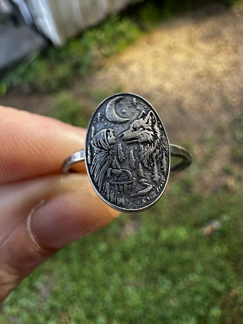 May include: Silver ring with an oval pendant featuring a detailed relief of a person facing a wolf under a crescent moon and stars. The ring has a thin band and a nature-inspired theme.