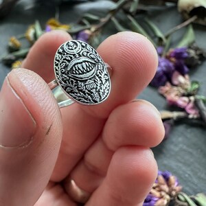 May include: Silver ring with an oval face featuring a detailed design of a mouth with sharp teeth, surrounded by ornate floral patterns. The ring is held up close, showcasing the intricate metalwork and gothic aesthetic.