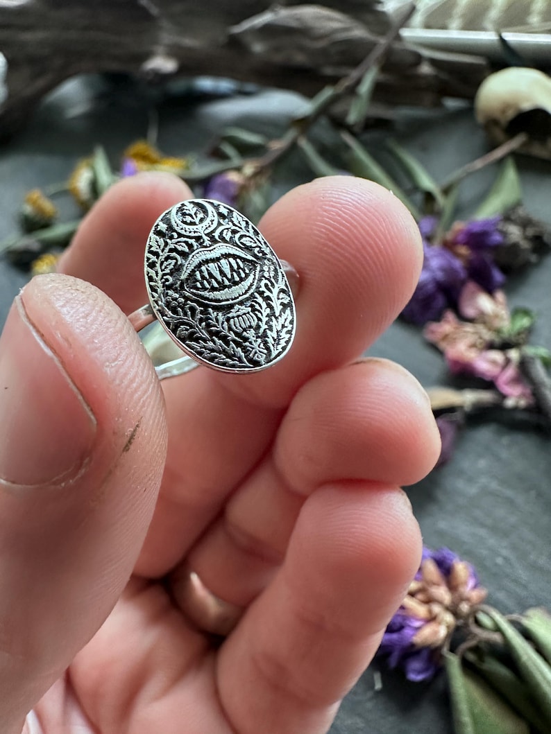 May include: Silver ring with an oval face featuring a detailed design of a mouth with sharp teeth, surrounded by ornate floral patterns. The ring is held up close, showcasing the intricate metalwork and gothic aesthetic.