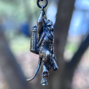 May include: A silver-toned pendant depicting a horned, goat-like figure carrying a basket on its back. The pendant is suspended from a small ring. The figure has a long beard and is detailed with intricate textures. The background is blurred, suggesting an outdoor setting.