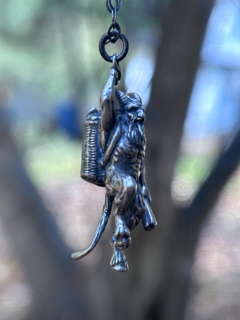 May include: A silver-toned pendant depicting a horned, goat-like figure carrying a basket on its back. The pendant is suspended from a small ring. The figure has a long beard and is detailed with intricate textures. The background is blurred, suggesting an outdoor setting.