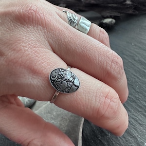 May include: Silver ring with an oval, engraved design featuring celestial and geometric patterns. The ring's band is thin and delicate, and the engraved design has a dark, oxidized finish. The ring is worn on a finger.