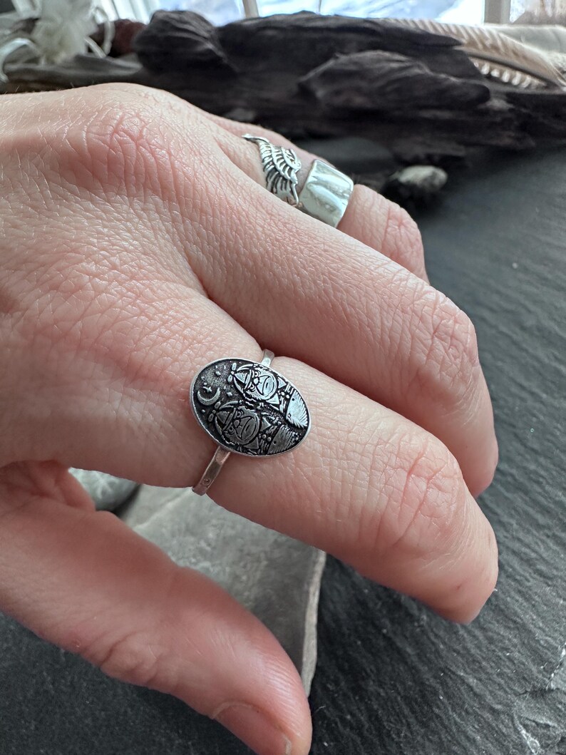 May include: Silver ring with an oval, engraved design featuring celestial and geometric patterns. The ring's band is thin and delicate, and the engraved design has a dark, oxidized finish. The ring is worn on a finger.