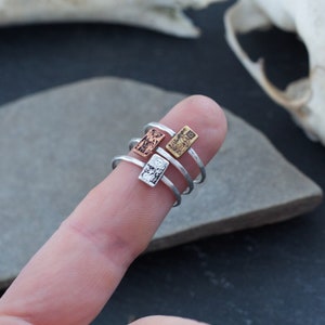 May include: Three silver rings with different textured rectangular charms. The charms are stacked on top of each other on a finger.