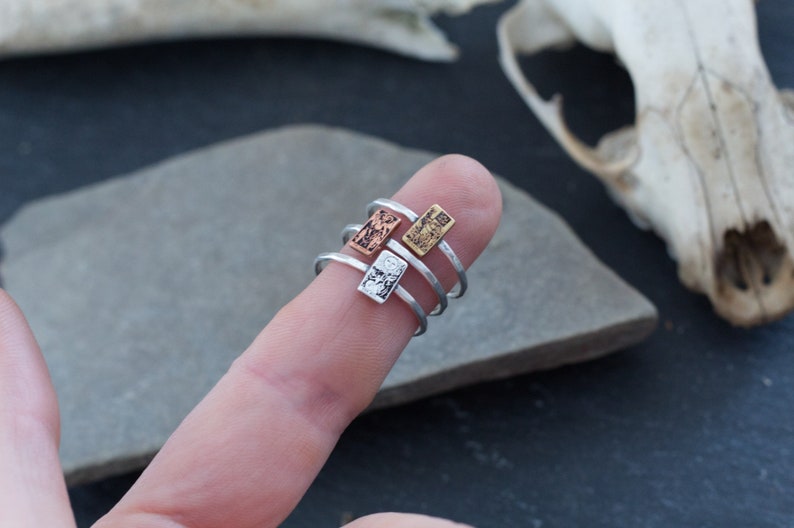 May include: Three silver rings with different textured rectangular charms. The charms are stacked on top of each other on a finger.