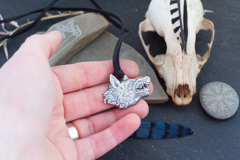 May include: Silver wolf head pendant necklace with a black cord. The pendant features a detailed wolf head with a circular design inside. The pendant is held in a hand with a ring on the finger.