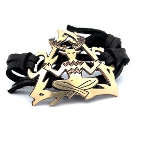 May include: A leather bracelet with a gold-toned metal charm featuring a stylized figure with antlers. The figure is sitting in a triangular shape.