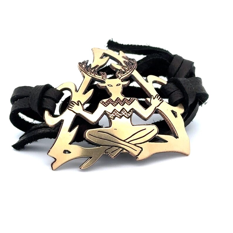May include: A leather bracelet with a gold-toned metal charm featuring a stylized figure with antlers. The figure is sitting in a triangular shape.