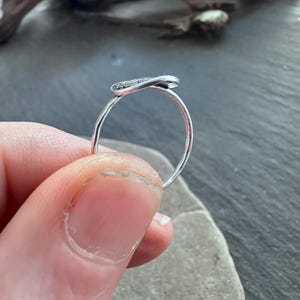 May include: A silver ring with a textured, curved design on top. The ring is held up against a dark background, showcasing its delicate details and minimalist style. The ring's band is thin and polished.