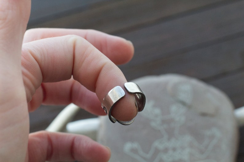 May include: A silver adjustable ring with a simple design. The ring is open on one side and can be adjusted to fit different finger sizes.