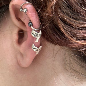 May include: A close-up of a person's ear with multiple ear piercings. The ear is adorned with a silver hoop earring with two silver beads, a silver cartilage earring with a black bead, and two silver ear cuffs with the words "NNX" and "MPT" inscribed on them.