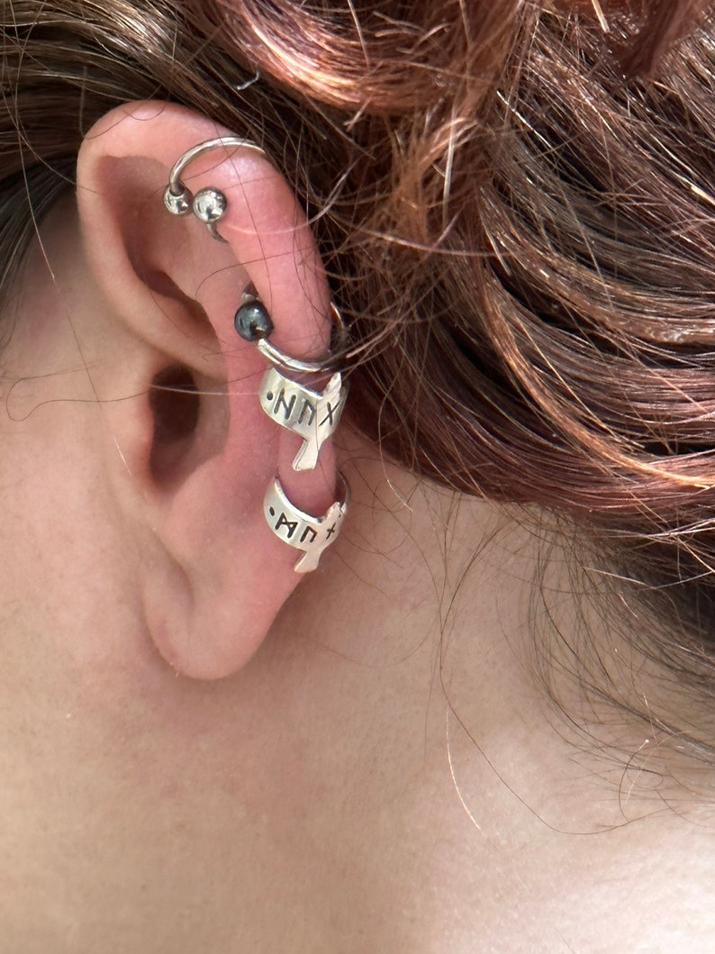 May include: A close-up of a person's ear with multiple ear piercings. The ear is adorned with a silver hoop earring with two silver beads, a silver cartilage earring with a black bead, and two silver ear cuffs with the words "NNX" and "MPT" inscribed on them.