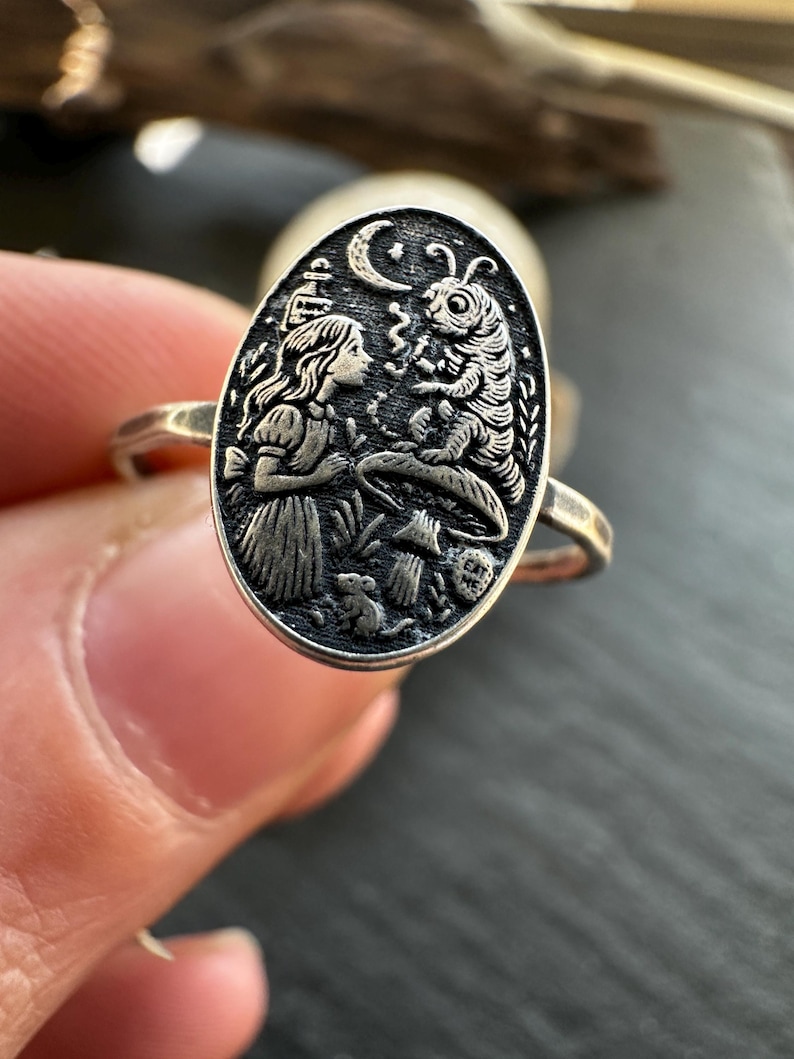 May include: Silver ring with an oval pendant featuring a detailed engraving. The design depicts a scene with a girl, a caterpillar, a crescent moon, and other elements. The ring is held by a person.