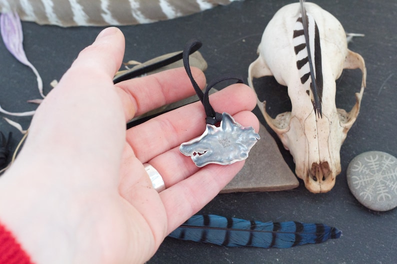 May include: A silver wolf head pendant on a black cord necklace. The pendant is detailed and textured, with a wolf's face in profile. The pendant is being held in a hand, with a blue and black feather and a small skull in the background.