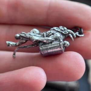 May include: A detailed silver-toned pendant featuring a skeletal figure riding a horned creature. The pendant has intricate detailing, with a purple-hued rectangular element at the base. The design suggests a fantasy or gothic theme, suitable for jewelry.