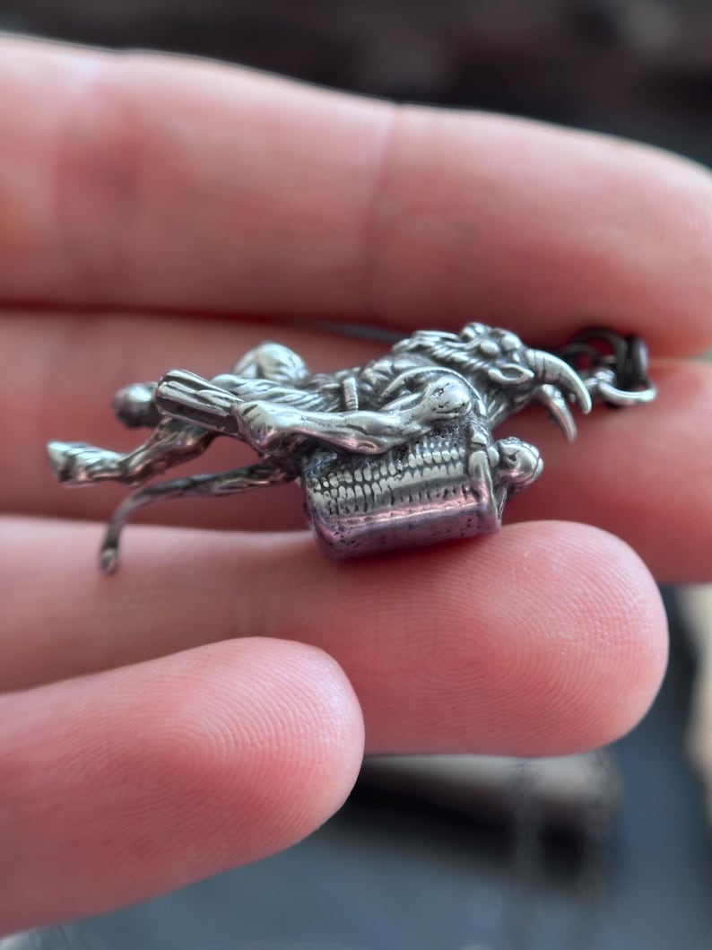 May include: A detailed silver-toned pendant featuring a skeletal figure riding a horned creature. The pendant has intricate detailing, with a purple-hued rectangular element at the base. The design suggests a fantasy or gothic theme, suitable for jewelry.