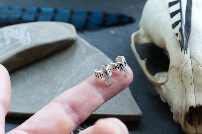 May include: A pair of silver hoop earrings with a bird design. The earrings are engraved with runes.