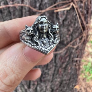 K&ouml;nnte beinhalten: Ein silberner Ring mit dem Gesicht einer Frau und zwei Katzen auf beiden Seiten. Der Ring hat detaillierte Merkmale und einen Vintage-Stil. Der Ring wird von einer Hand gehalten.