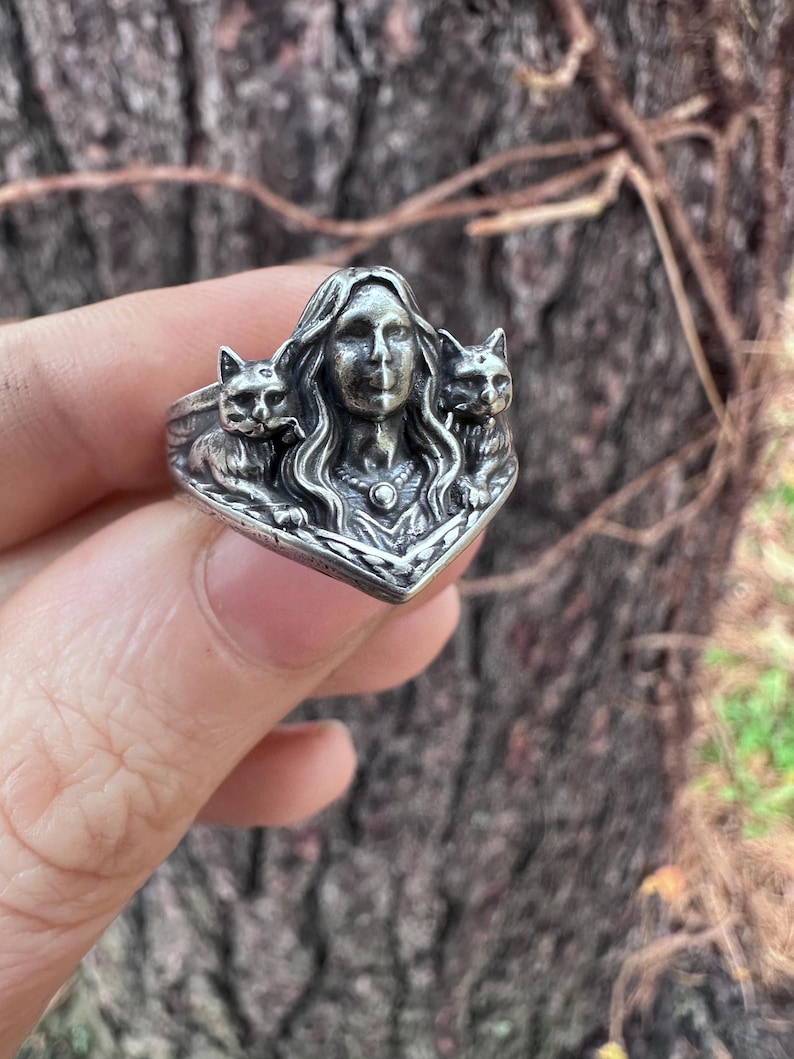 K&ouml;nnte beinhalten: Ein silberner Ring mit dem Gesicht einer Frau und zwei Katzen auf beiden Seiten. Der Ring hat detaillierte Merkmale und einen Vintage-Stil. Der Ring wird von einer Hand gehalten.