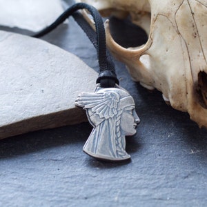 May include: A silver pendant necklace with a detailed profile of a woman's face with a feathered headdress. The necklace is on a black leather cord and is resting on a gray stone surface next to a white animal skull.