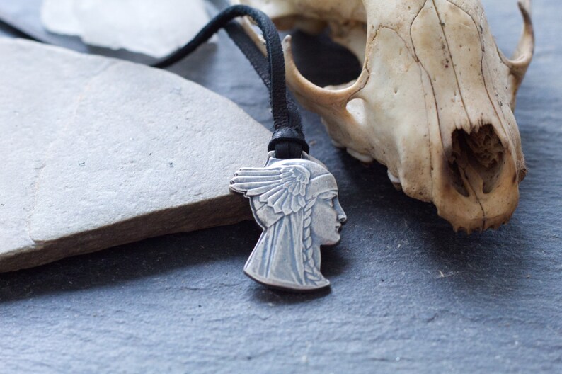 May include: A silver pendant necklace with a detailed profile of a woman's face with a feathered headdress. The necklace is on a black leather cord and is resting on a gray stone surface next to a white animal skull.