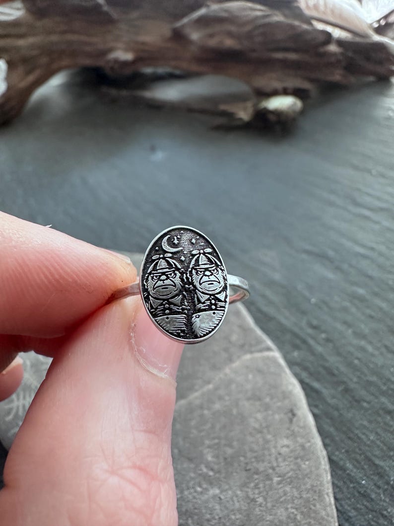 May include: Silver ring featuring an oval-shaped face with two figures under a crescent moon and stars. The ring has a detailed design with a vintage or antique aesthetic. The ring is held by a person's fingers.