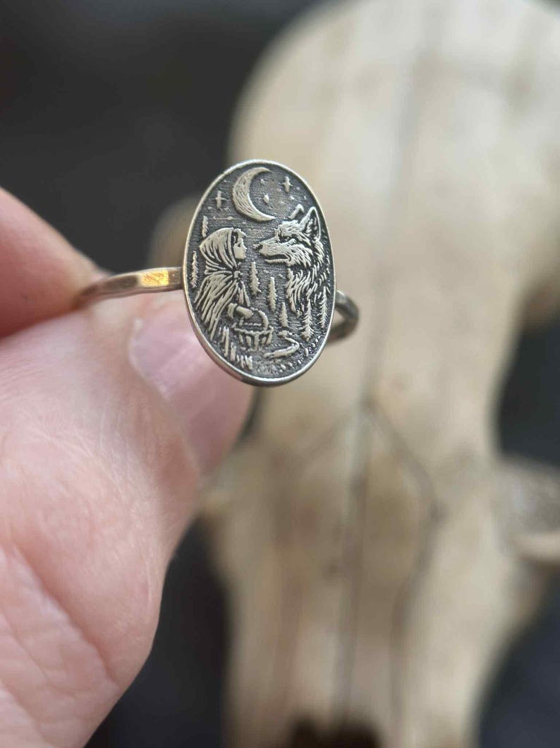 May include: Silver-toned ring with an oval pendant featuring an engraved scene of a girl and a wolf under a crescent moon. The ring's design includes trees and a basket, creating a fairytale theme. The ring is held by a hand.
