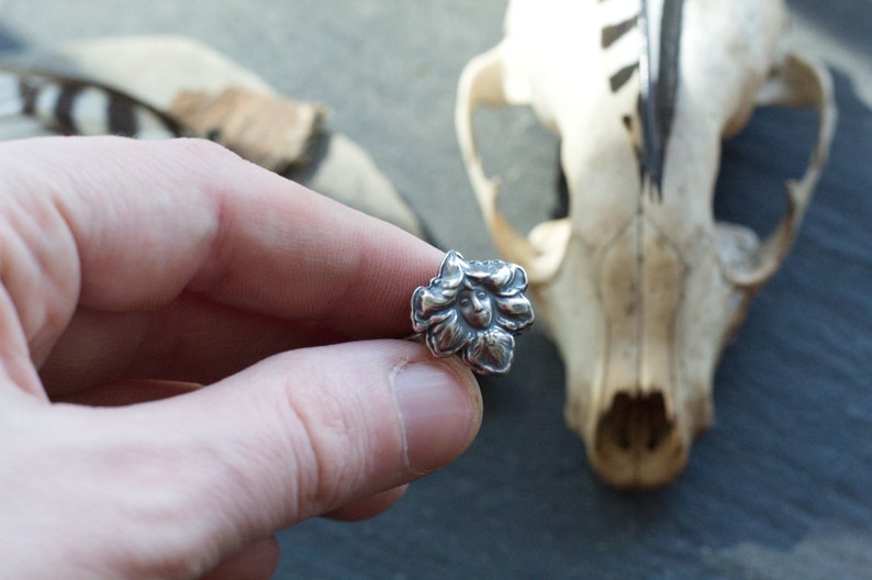 May include: A silver ring featuring a detailed flower design. The flower has a face in the centre.