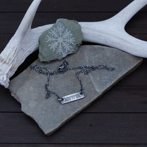 May include: A silver chain necklace with a silver bar pendant engraved with the word "DOMINIX". The necklace is displayed on a grey stone with a white antler behind it.