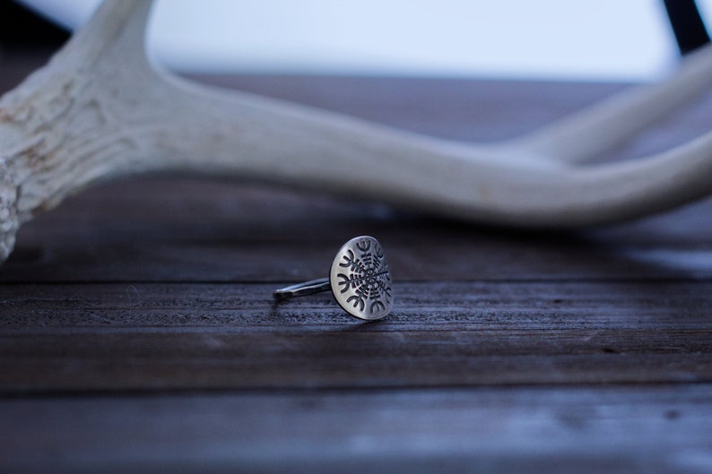 May include: A silver ring with a circular design featuring a snowflake-like pattern. The ring is lying on a wooden surface.