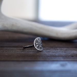 May include: A silver ring with a circular design featuring a compass rose with eight points. The ring is on a dark brown wooden surface.