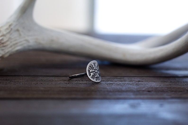 May include: A silver ring with a circular design featuring a compass rose with eight points. The ring is on a dark brown wooden surface.