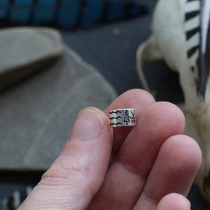 May include: A silver ring with a design of three trees and branches. The ring is being held by a hand.