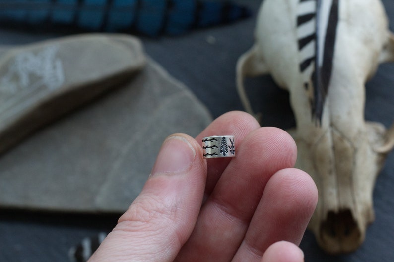 May include: A silver ring with a design of three trees and branches. The ring is being held by a hand.