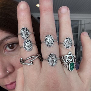 May include: A hand displays several silver rings with various designs, including celestial motifs, a tree, and a star. One ring features a green gemstone. The rings are oval-shaped and have intricate details.