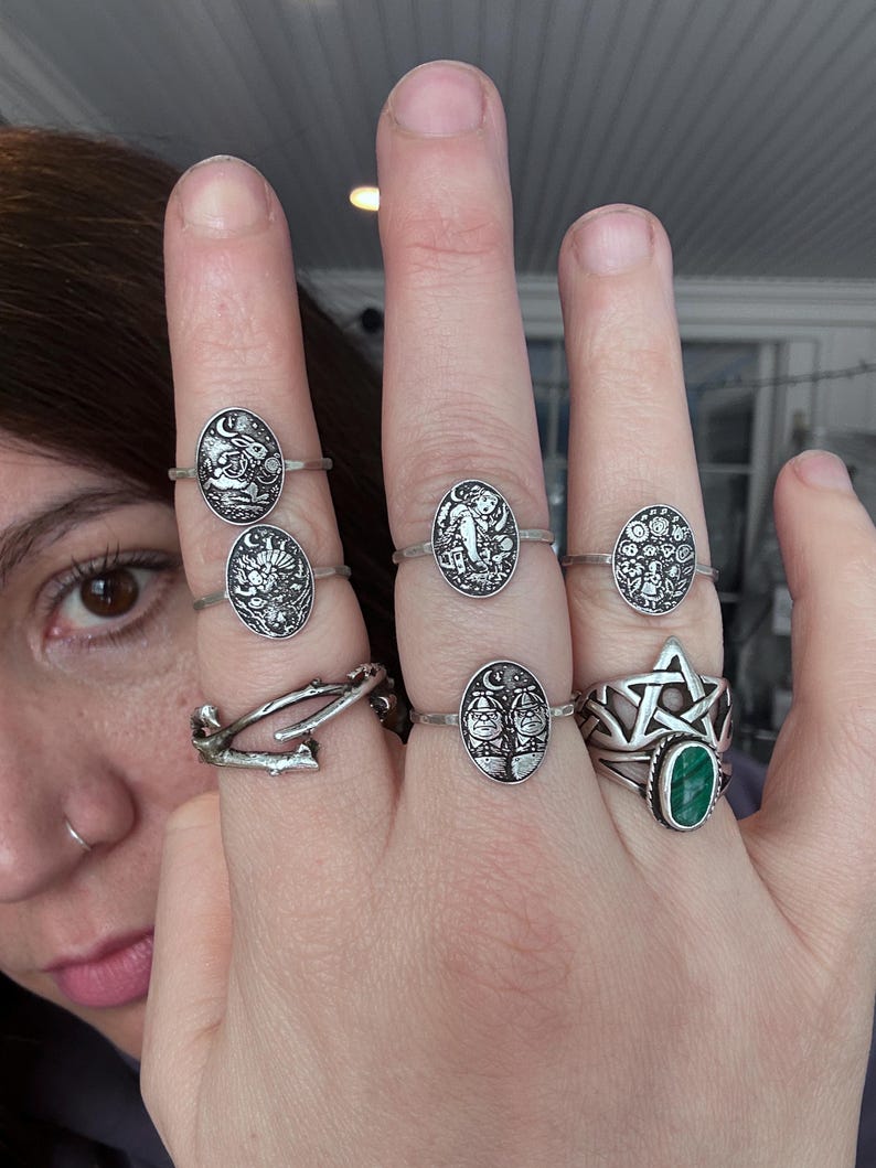 May include: A hand displays several silver rings with various designs, including celestial motifs, a tree, and a star. One ring features a green gemstone. The rings are oval-shaped and have intricate details.
