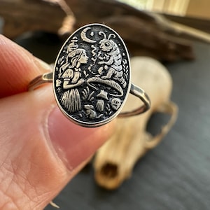May include: Silver ring with an oval pendant featuring an engraved scene from Alice in Wonderland. The design includes Alice, a caterpillar, mushrooms, and a crescent moon. The ring is held up by a finger.