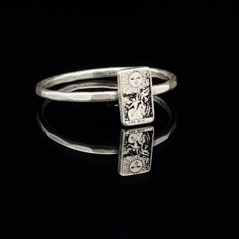 May include: A silver ring with a small rectangular charm featuring a tarot card design. The card depicts the Sun card with the text "The Sun" below the image.