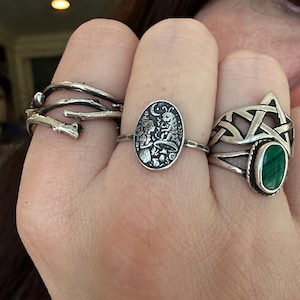 May include: A hand displays three silver rings. One ring has a twig-like design, another features an oval with a detailed forest scene, and the third has a green stone set within a Celtic knot design.