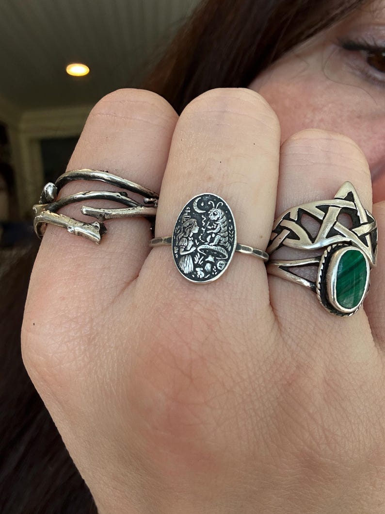 May include: A hand displays three silver rings. One ring has a twig-like design, another features an oval with a detailed forest scene, and the third has a green stone set within a Celtic knot design.