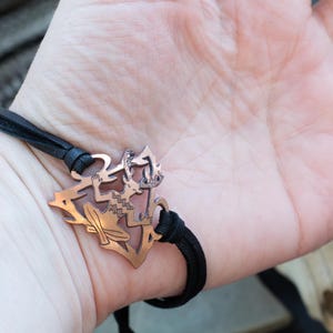 May include: A black leather bracelet with a copper charm featuring a stylized figure of a person with wings. The charm is detailed with intricate patterns.