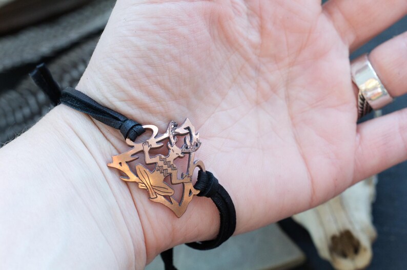 May include: A black leather bracelet with a copper charm featuring a stylized figure of a person with wings. The charm is detailed with intricate patterns.