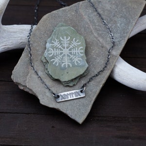 May include: A silver chain necklace with a silver bar pendant engraved with the Roman numeral MCMXCIX. The necklace is displayed on a grey stone with a white etched symbol.