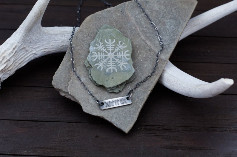 May include: A silver chain necklace with a silver bar pendant engraved with the Roman numeral MCMXCIX. The necklace is displayed on a grey stone with a white etched symbol.