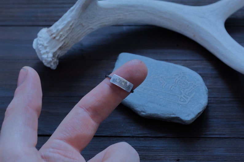 May include: A silver ring with a rectangular face featuring a detailed landscape design. The ring is worn on a finger, set against a dark wood surface with a stone and a white antler in the background. The ring's band is thin.
